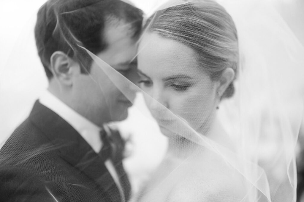 A close-up of a couple wrapped in veils, sharing a tender moment, captured in soft black and white.