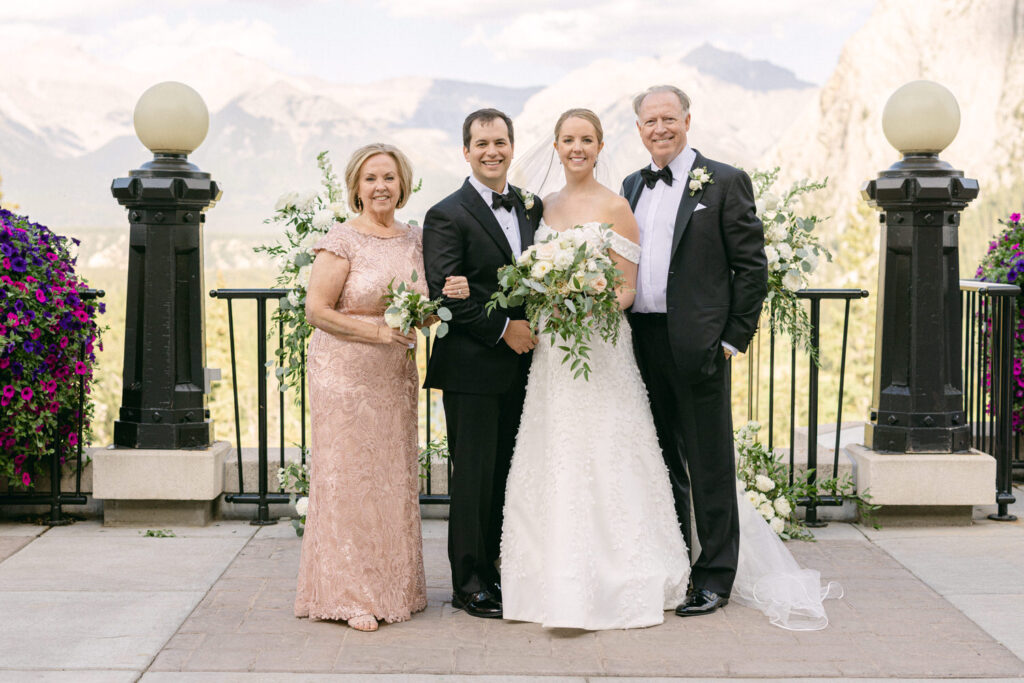 A joyful family poses together at a wedding, featuring the bride in a beautiful white gown, flanked by her parents and a relative amidst vibrant flowers and majestic mountains in the background.