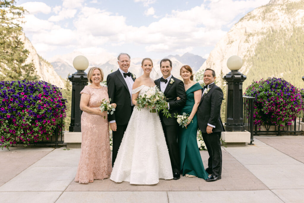 A joyful bride and groom pose with family members against a backdrop of vibrant flowers and majestic mountains on a beautiful day.