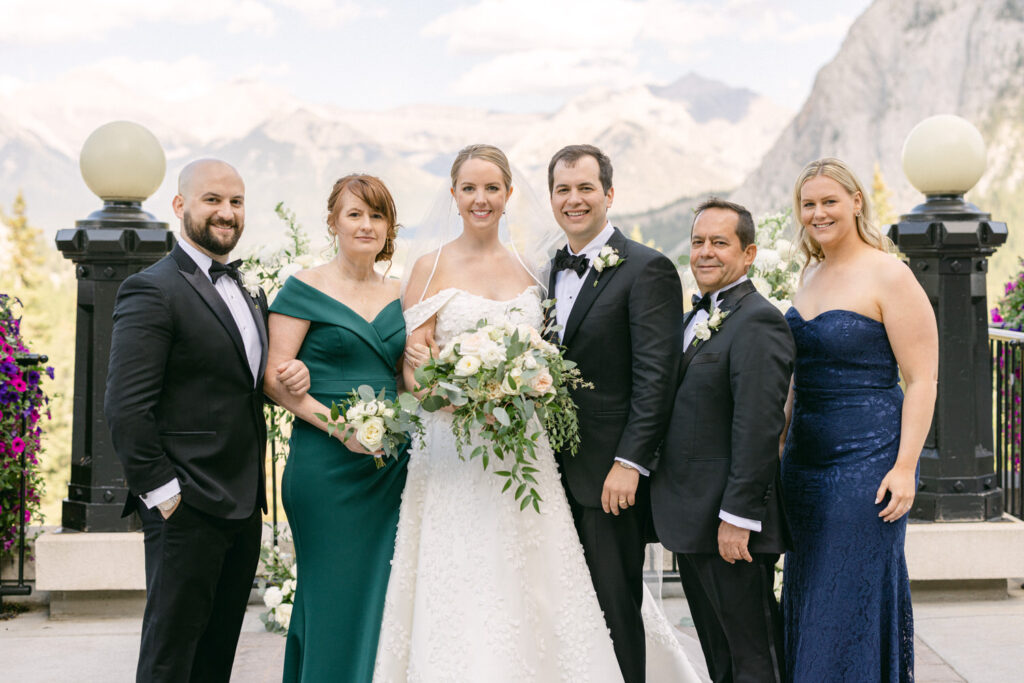 A joyful wedding party posing for a photo outdoors, surrounded by beautiful flowers and mountains in the background.