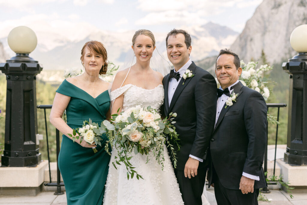 A joyful wedding couple poses with their parents against a backdrop of mountains and greenery, showcasing their elegant attire and floral arrangements.