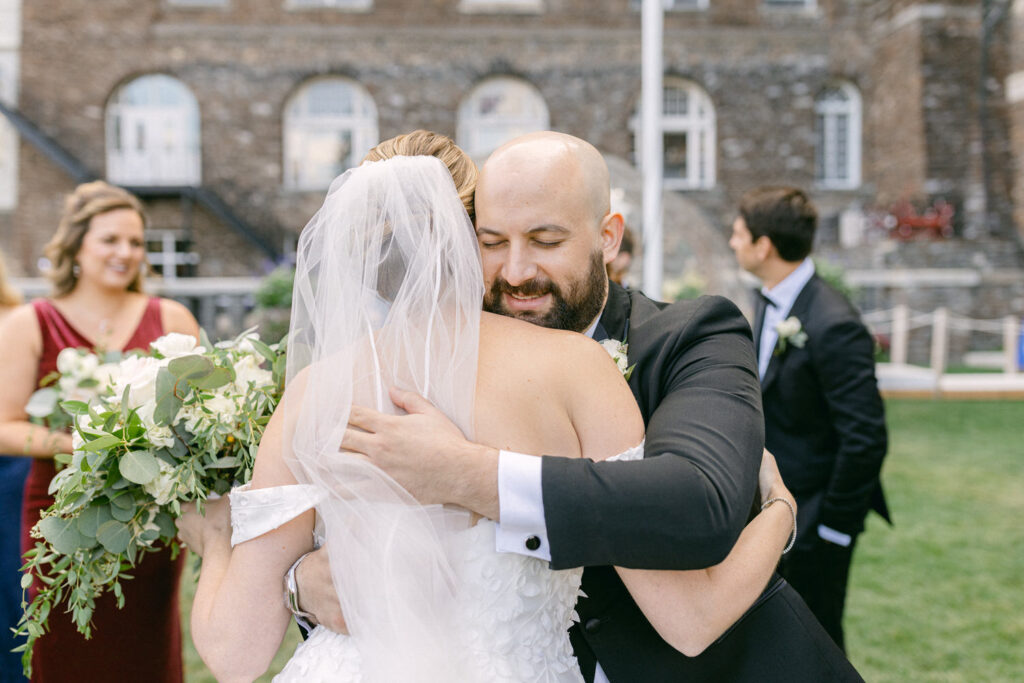 A groom warmly hugs his bride, both radiating happiness and love, surrounded by wedding guests and greenery.