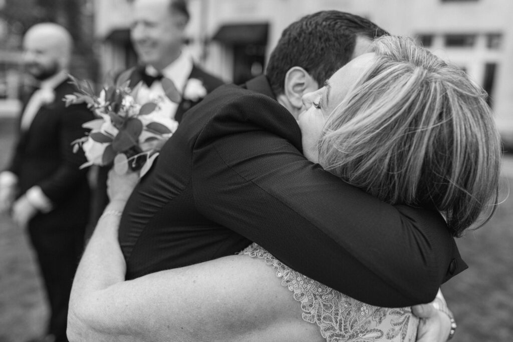 A joyful hug between two people dressed elegantly, with floral arrangements in the foreground and smiling guests in the background.