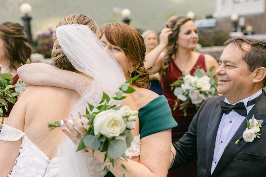 A joyful embrace between a bride and her mother, surrounded by bridesmaids and family, with floral arrangements in hand and a picturesque backdrop.