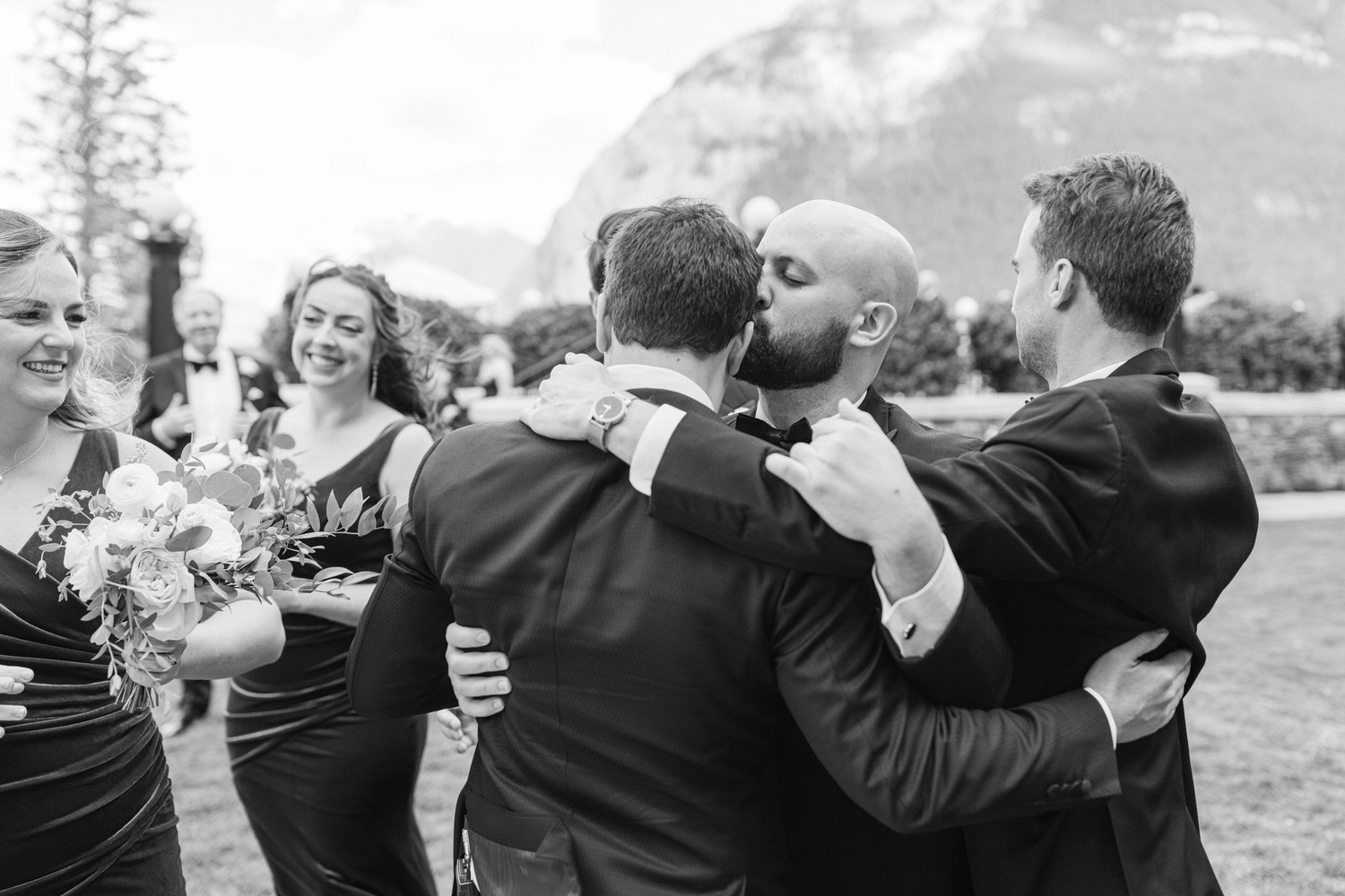 A joyful group of wedding guests sharing hugs and laughter outdoors, with mountains in the background.