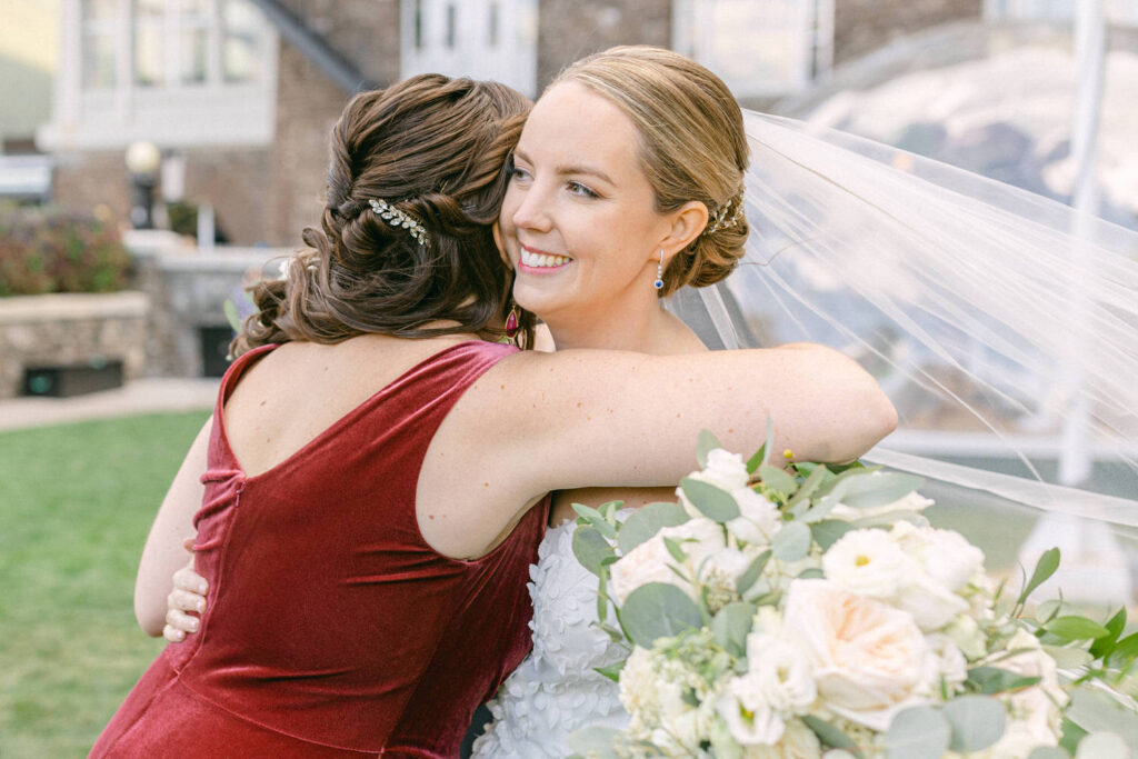 A bride and her friend share a joyful hug, surrounded by greenery and soft floral arrangements, capturing a moment of celebration and friendship.