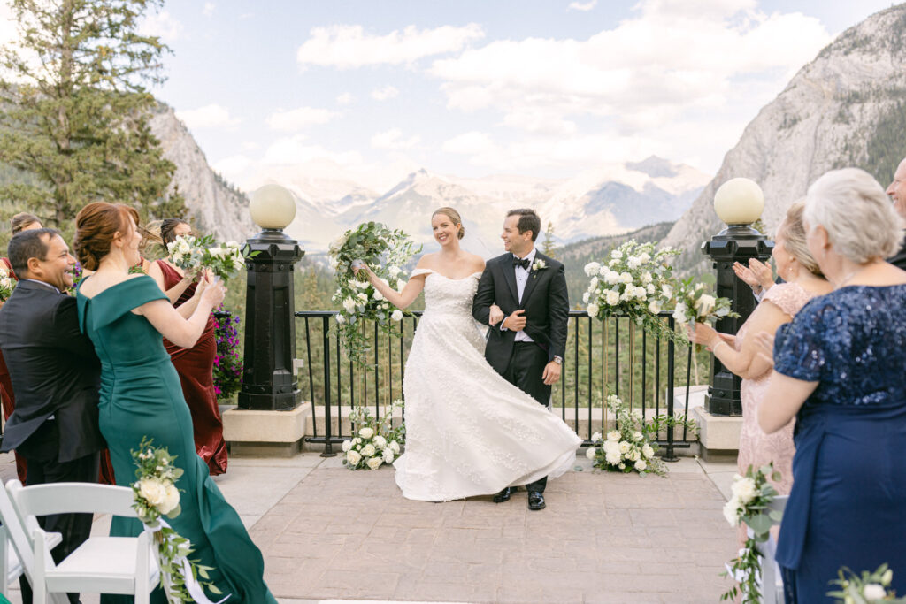 A joyful couple exits their wedding ceremony, surrounded by guests and stunning mountain scenery.