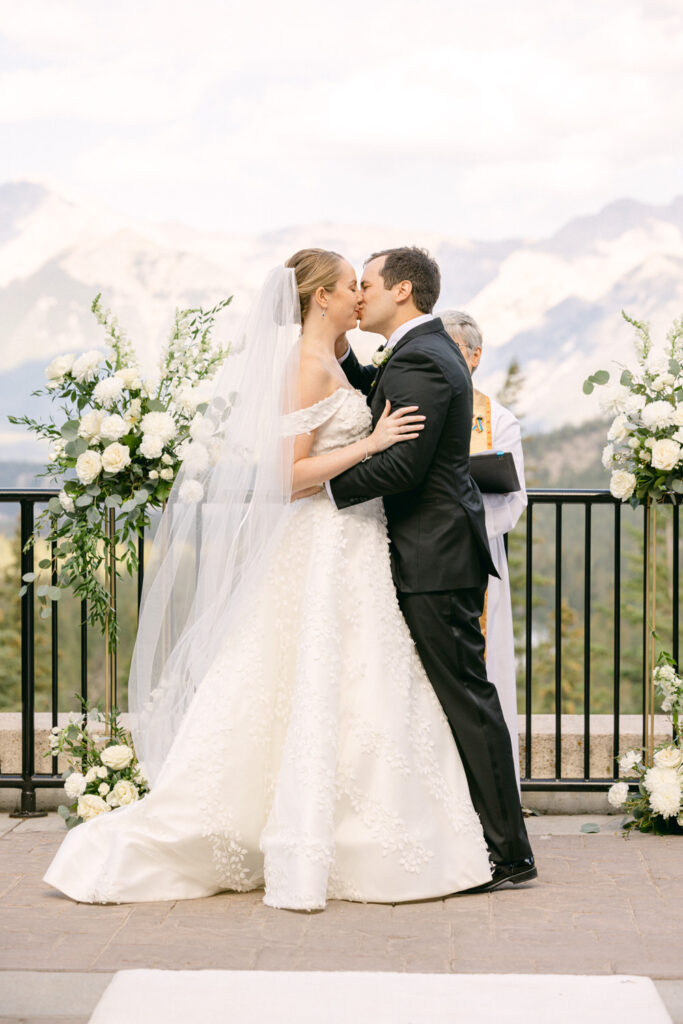 A newlywed couple shares a kiss during their outdoor wedding ceremony with mountains in the background and floral arrangements nearby.