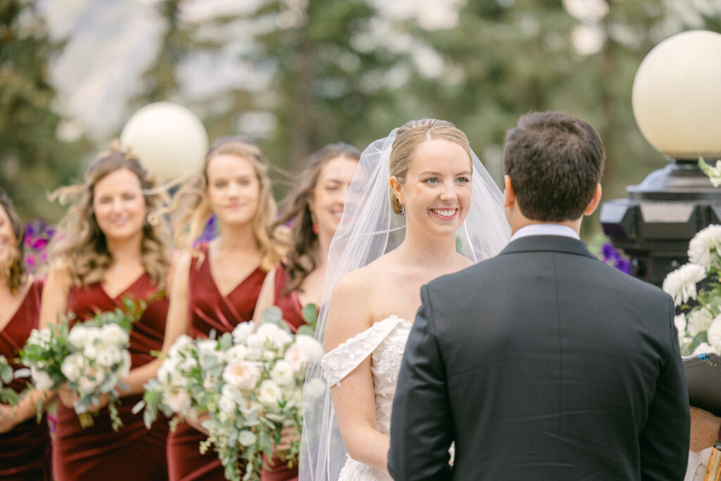 A bride smiles joyfully as she exchanges vows with her partner, surrounded by her bridesmaids in burgundy dresses and floral arrangements.