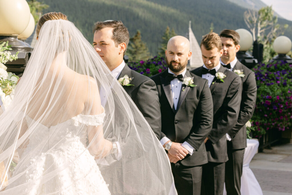 A bride with a veil stands facing her partner during a wedding ceremony, surrounded by groomsmen in tuxedos against a scenic mountain backdrop.