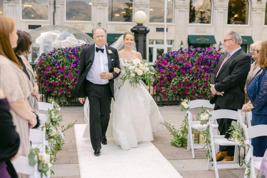 A bride in a white gown, accompanied by her father in a tuxedo, walks down a flower-adorned aisle amid seated guests.