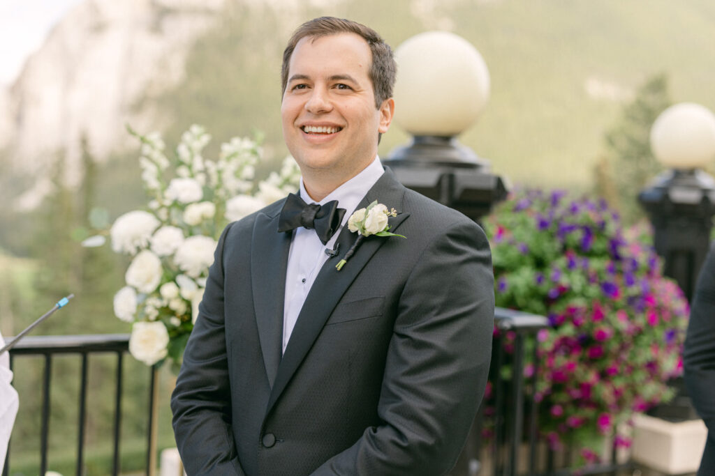 A cheerful groom in a black tuxedo stands outdoors, surrounded by floral arrangements and a scenic mountainous backdrop.