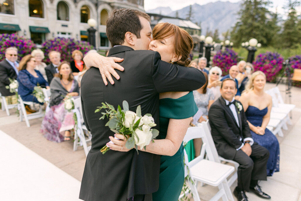 A joyous moment of a groom embracing his mother during an outdoor wedding ceremony, surrounded by guests and vibrant floral arrangements.