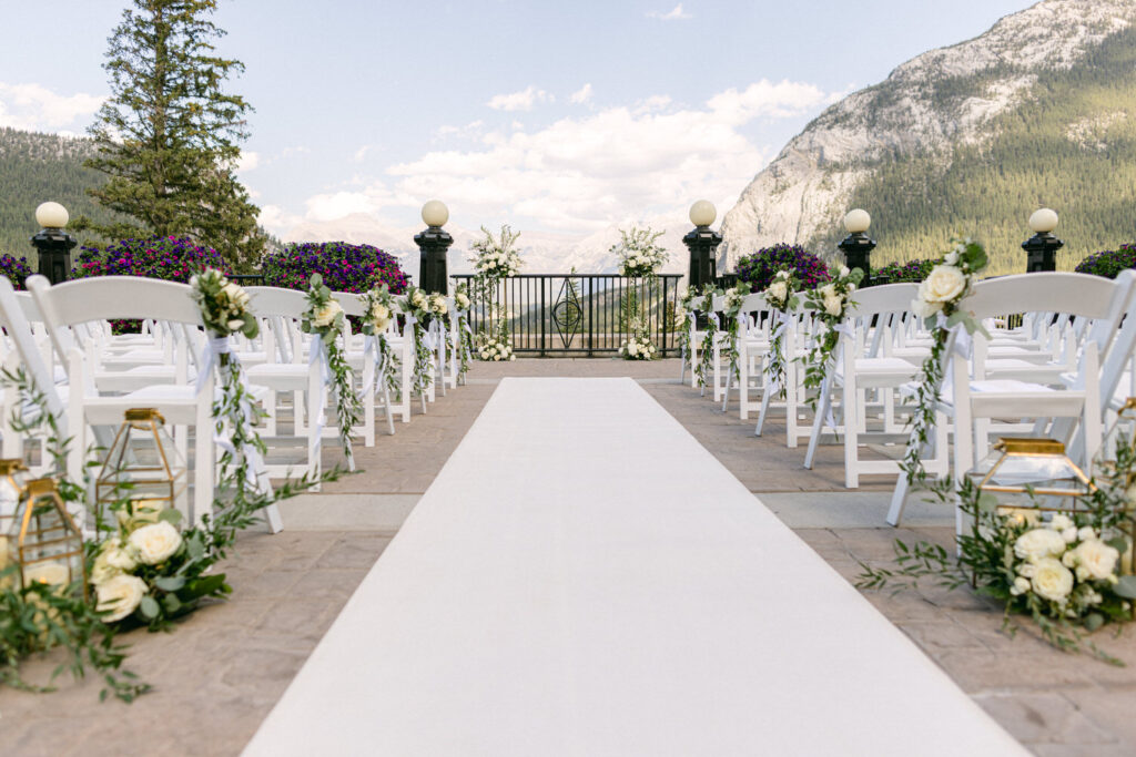 A beautifully arranged outdoor wedding aisle lined with white chairs adorned with floral decorations, leading to a scenic backdrop of mountains and clouds.