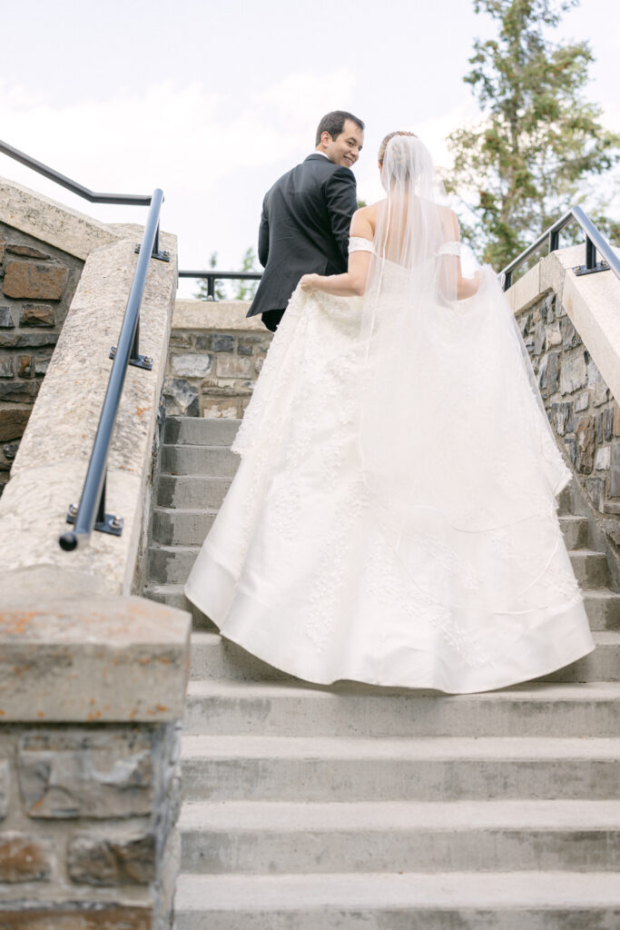 A bride and groom walk up a stone staircase, with the bride holding her flowing gown, both smiling at each other against a backdrop of trees and a blue sky.