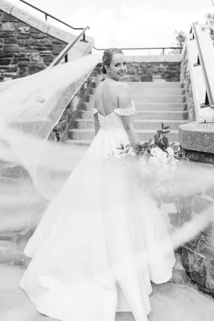 A bride in a stunning off-shoulder gown, holding a bouquet, smiles back as she walks up a stone staircase with her flowing veil trailing behind her.