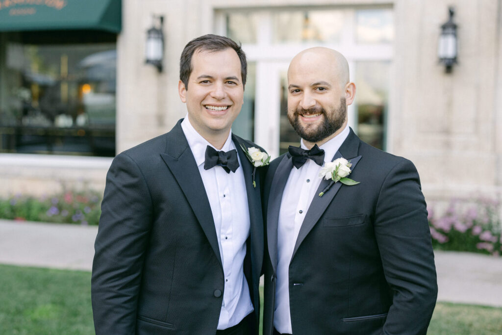 A joyful photo of two men in tuxedos, smiling together outdoors, each wearing a boutonniere, with a blurred elegant building and flowers in the background.