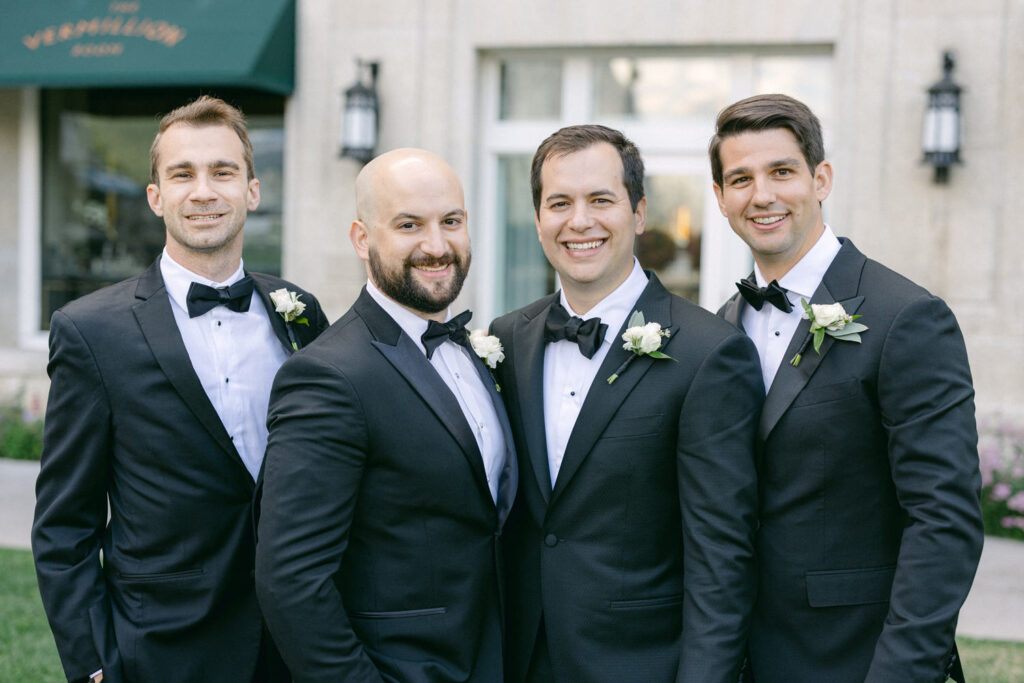 Group of four men in tuxedos smiling, each wearing a white flower boutonnière, standing outdoors with a venue in the background.