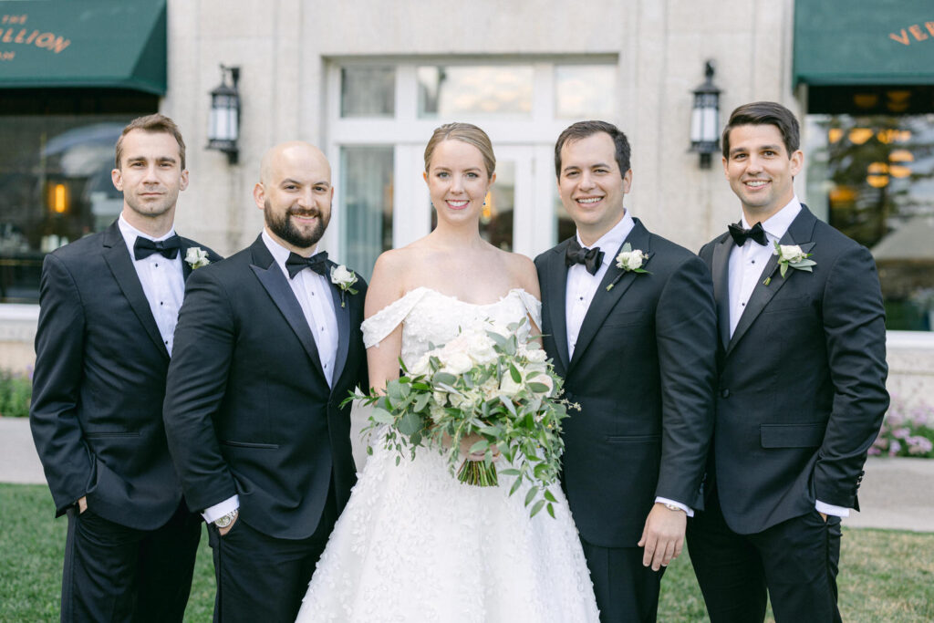 A bride in a strapless wedding gown holds a bouquet while posing with four groomsmen in tuxedos, outdoors in front of a beautiful venue.