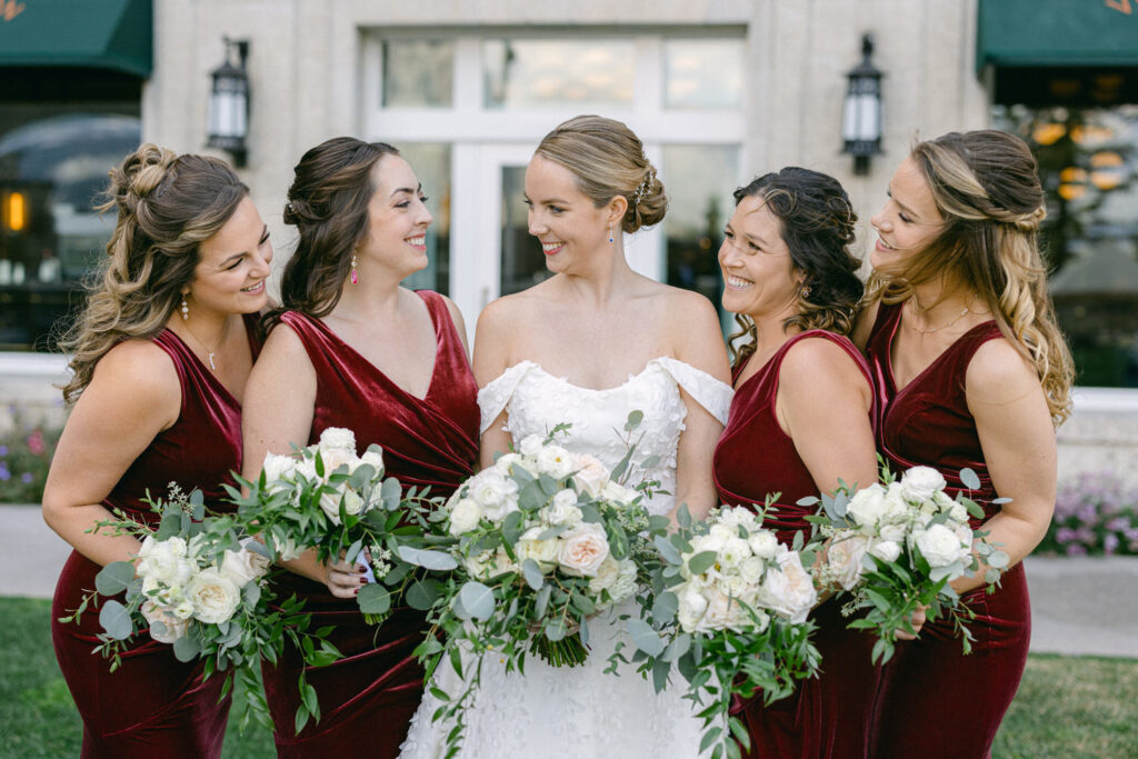 A smiling bride surrounded by her bridesmaids in burgundy dresses, each holding beautiful floral bouquets.