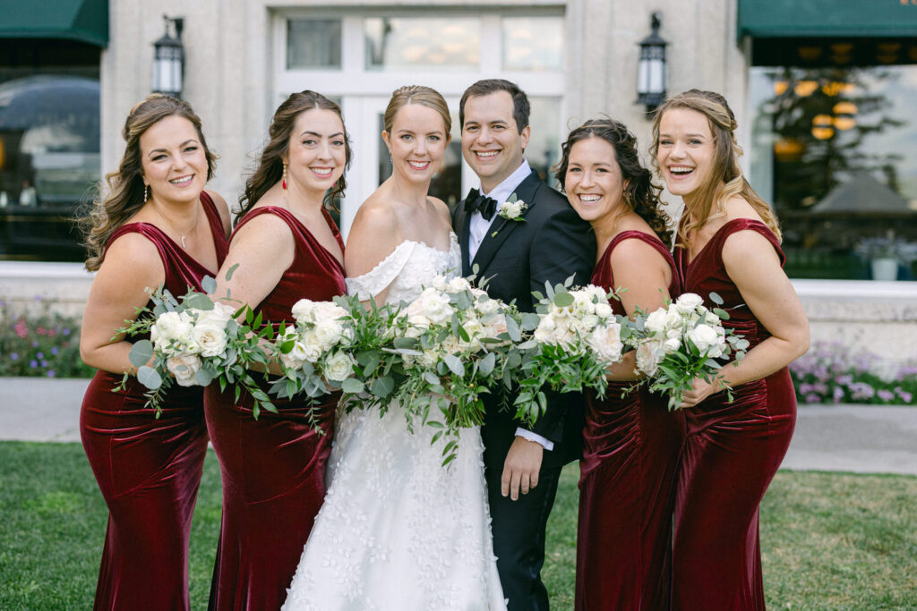 A joyful bride stands with her bridesmaids, all holding floral bouquets and dressed in elegant burgundy gowns, with a backdrop of greenery and a stylish venue.