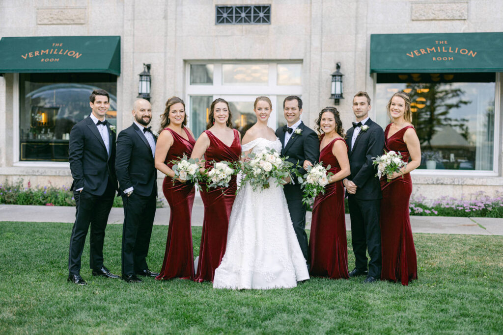 A joyful bridal party posing together outside The Vermillion Room, showcasing elegant burgundy dresses and a stunning bride in a white gown, surrounded by floral arrangements.