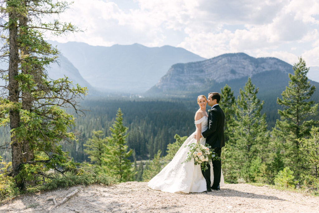 A newlywed couple embraces amidst a stunning mountain backdrop, surrounded by lush greenery and majestic peaks.