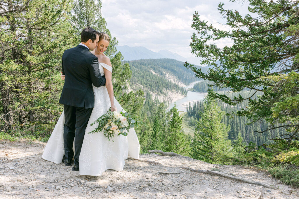 Couple Embracing in Nature::A bride and groom share a romantic moment in a scenic outdoor setting, surrounded by lush greenery and a river in the background.