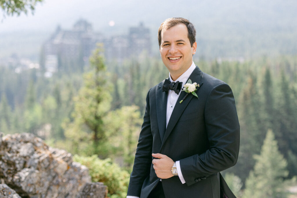 Elegant Groom in Nature::A smiling groom in a black tuxedo with a bow tie and floral boutonniere, standing against a backdrop of trees and a distant building.