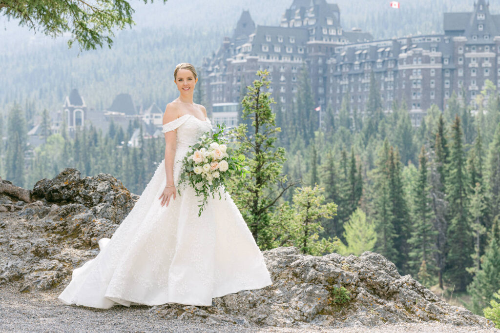 A smiling bride in an elegant off-the-shoulder wedding gown, holding a bouquet of flowers, poses in front of a stunning mountainous backdrop with a historic hotel surrounded by lush trees.