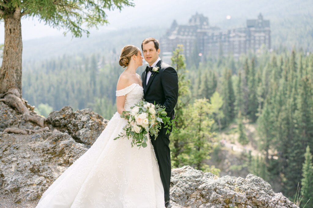 A couple stands together on a rocky ledge overlooking a lush green forest and a historic hotel in the background, captured in a beautiful outdoor setting.