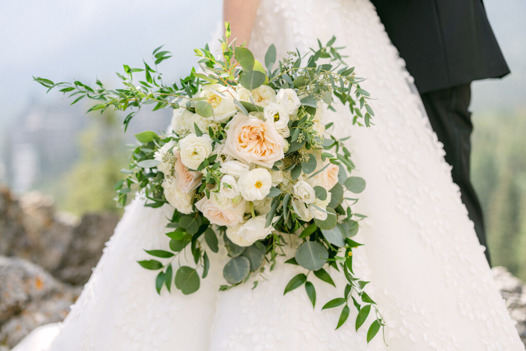 A close-up of a bride holding a lush bouquet featuring white and peach roses, accented with greenery, set against a scenic outdoor backdrop.