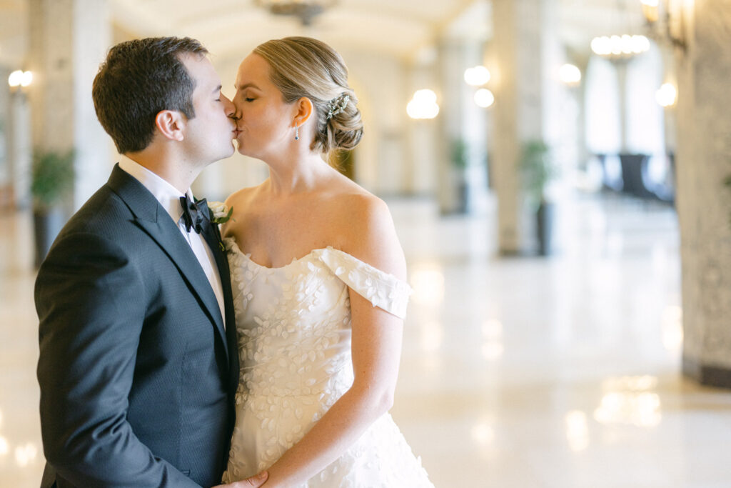 A newlywed couple shares a tender kiss in an elegant indoor setting, surrounded by soft lighting and decorative plants.