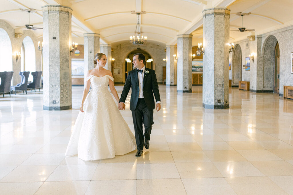 A couple in formal attire walks hand-in-hand through a beautifully decorated hallway with elegant lighting and architecture.