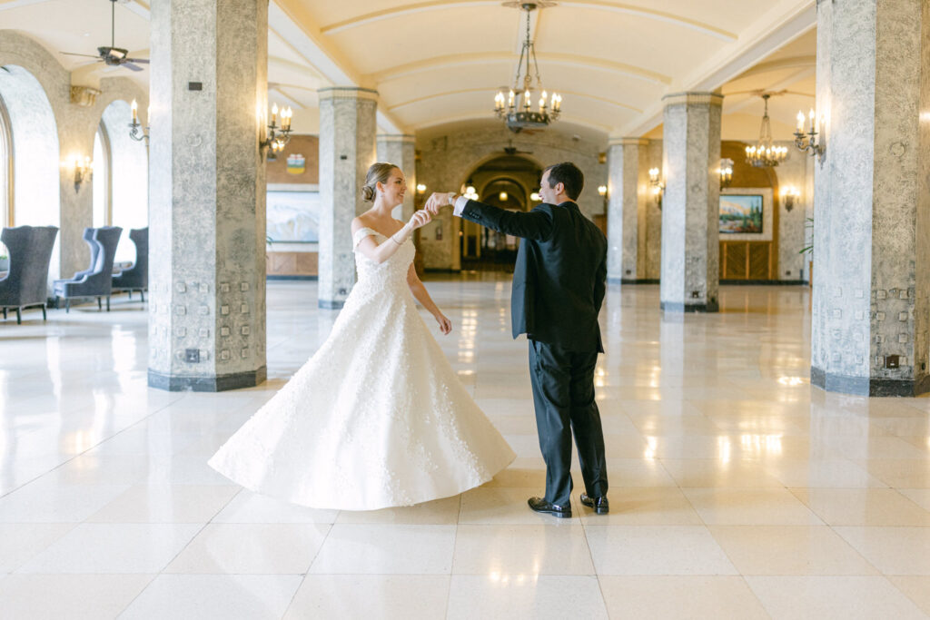 A bride and groom dance together in a beautifully lit, spacious hall with classic architecture and decorative chandeliers.