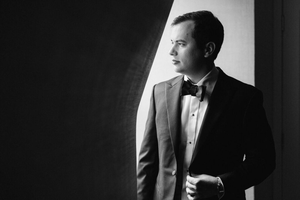 A man in a black tuxedo and bow tie stands by a window, gazing thoughtfully outside in a monochrome setting.