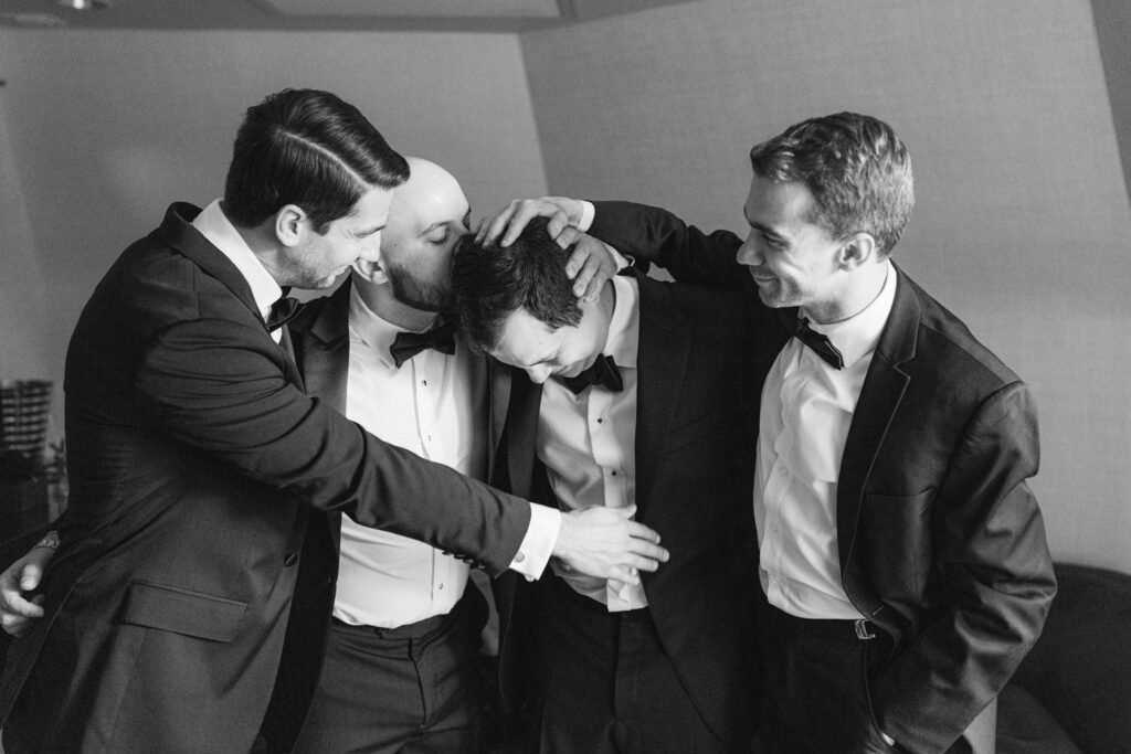 A group of four groomsmen in tuxedos share a joyful moment, showcasing camaraderie and laughter before a wedding ceremony.