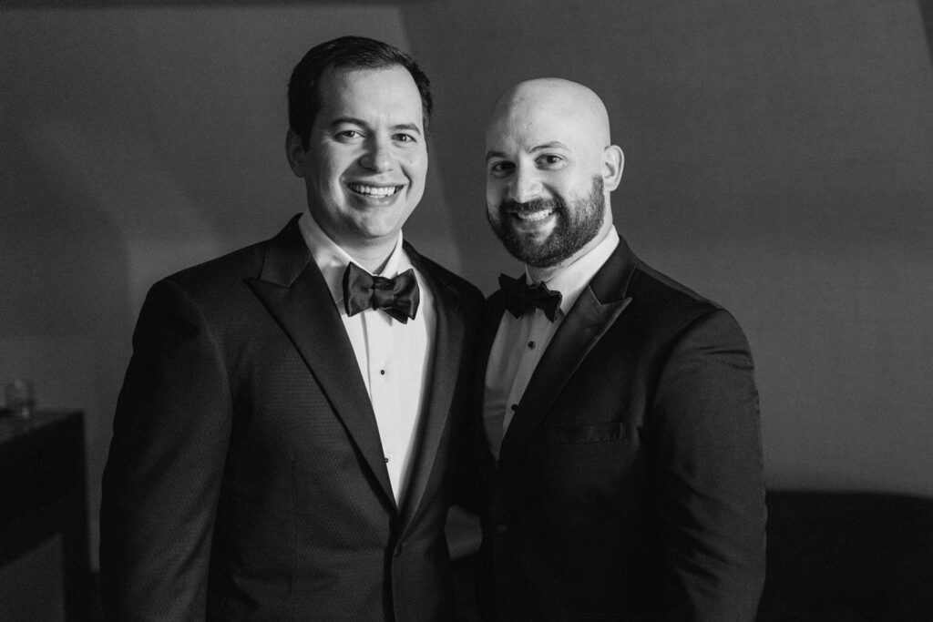 Two men in tuxedos smiling happily together in a softly lit environment.