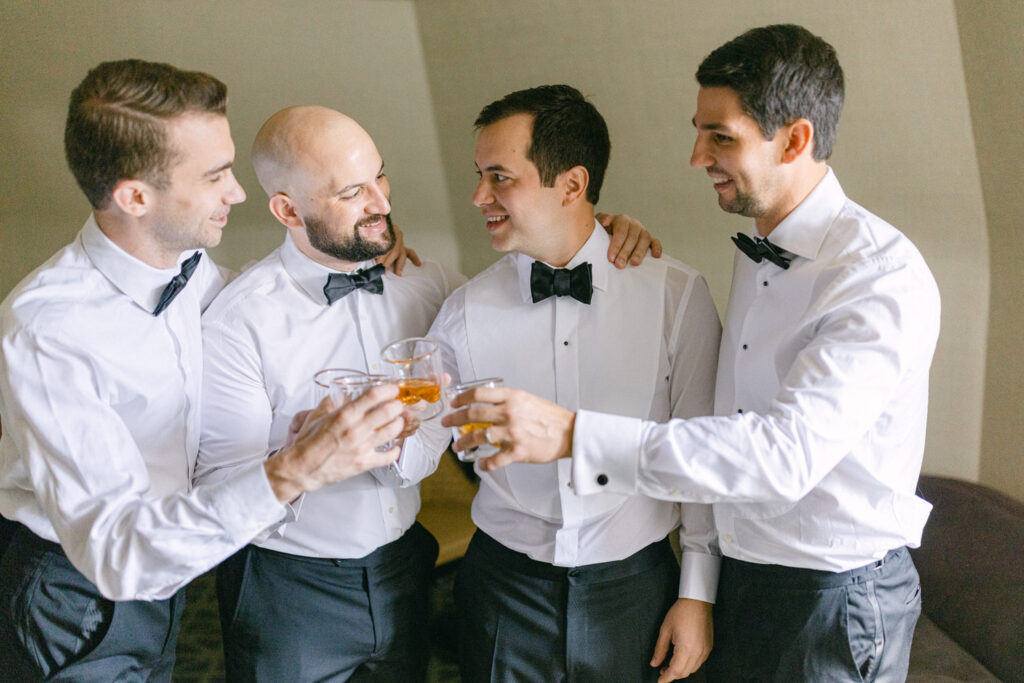 Four well-dressed groomsmen in tuxedos raise glasses in a toast, smiling and enjoying a moment of camaraderie before the wedding.