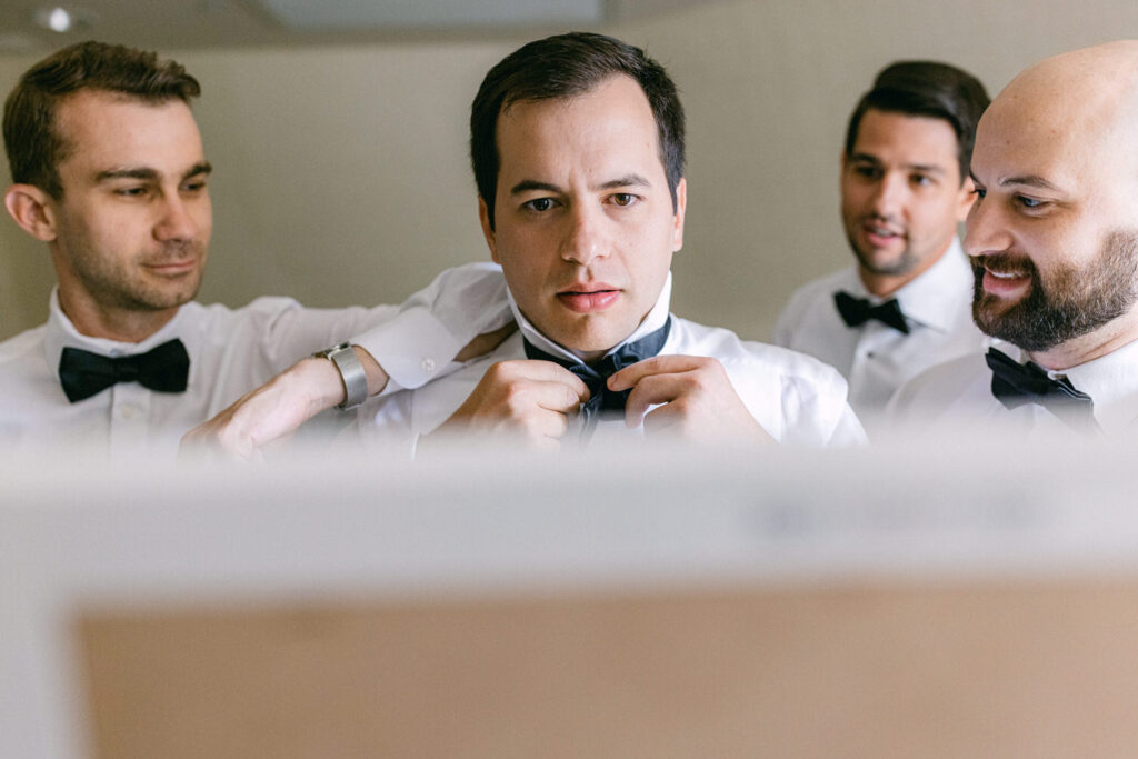 A groom is adjusting his bow tie with the help of friends, all dressed in white shirts and black bow ties, ready for a wedding.