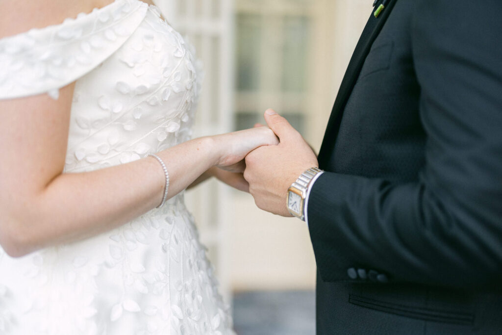 A couple holding hands during a romantic wedding ceremony, showcasing the bride's detailed dress and the groom's formal attire.