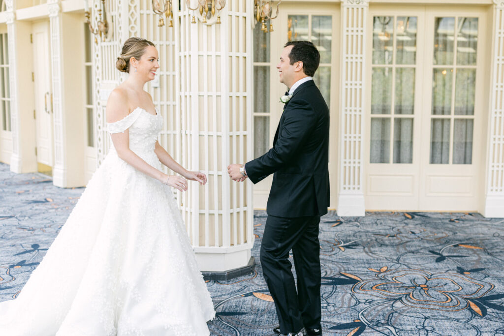 A bride in a beautiful white gown happily approaches a groom in a black tuxedo, both smiling in an elegant venue.