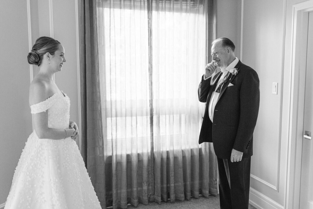 A bride in a beautiful off-the-shoulder wedding gown shares a heartfelt moment with her father, who is visibly moved, both smiling in an intimate setting with natural light filtering through sheer curtains.