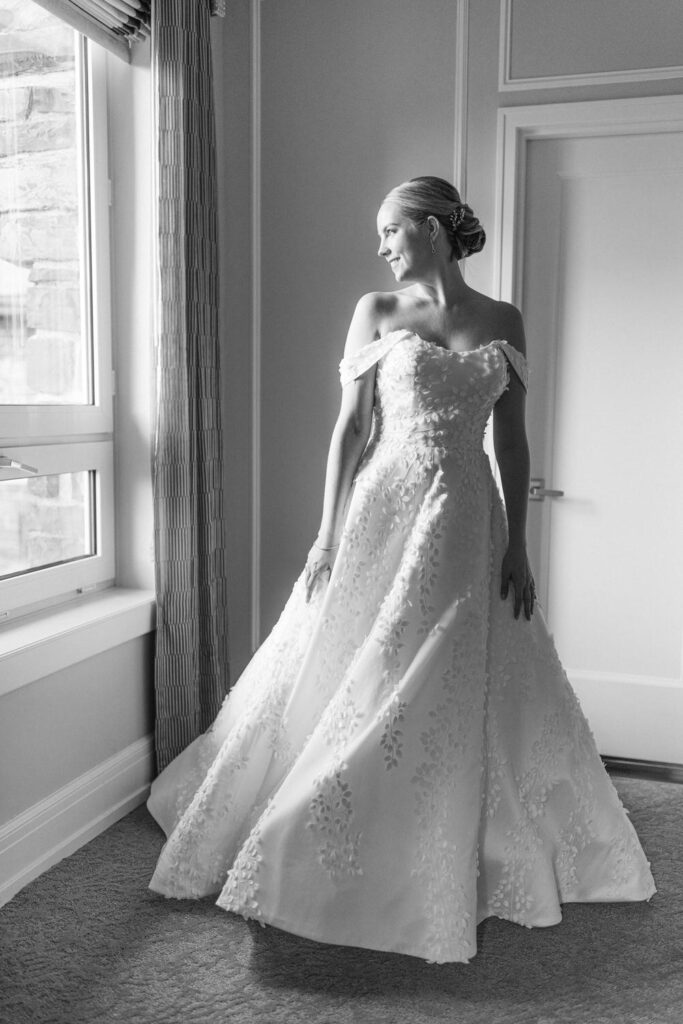 A smiling bride in an intricately adorned off-shoulder wedding gown, standing gracefully by a window with natural light illuminating the scene.