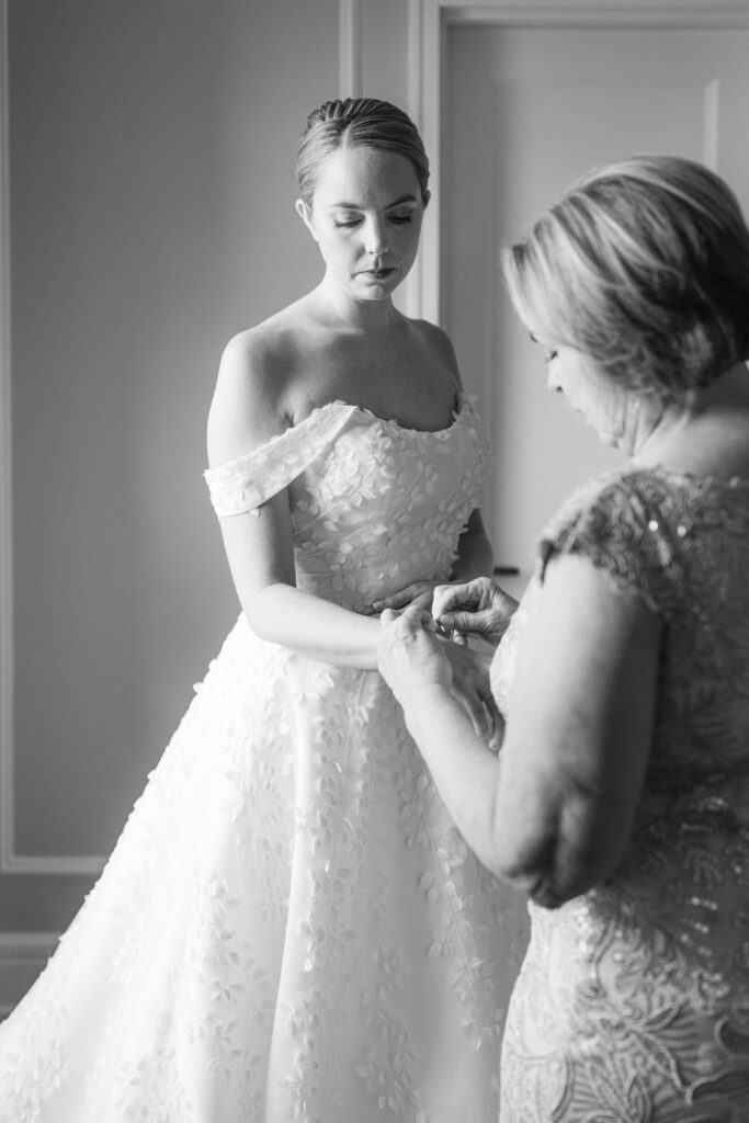 A bride in a beautiful off-the-shoulder wedding dress looks down as her mother assists her with a bracelet, capturing a tender moment before the ceremony.