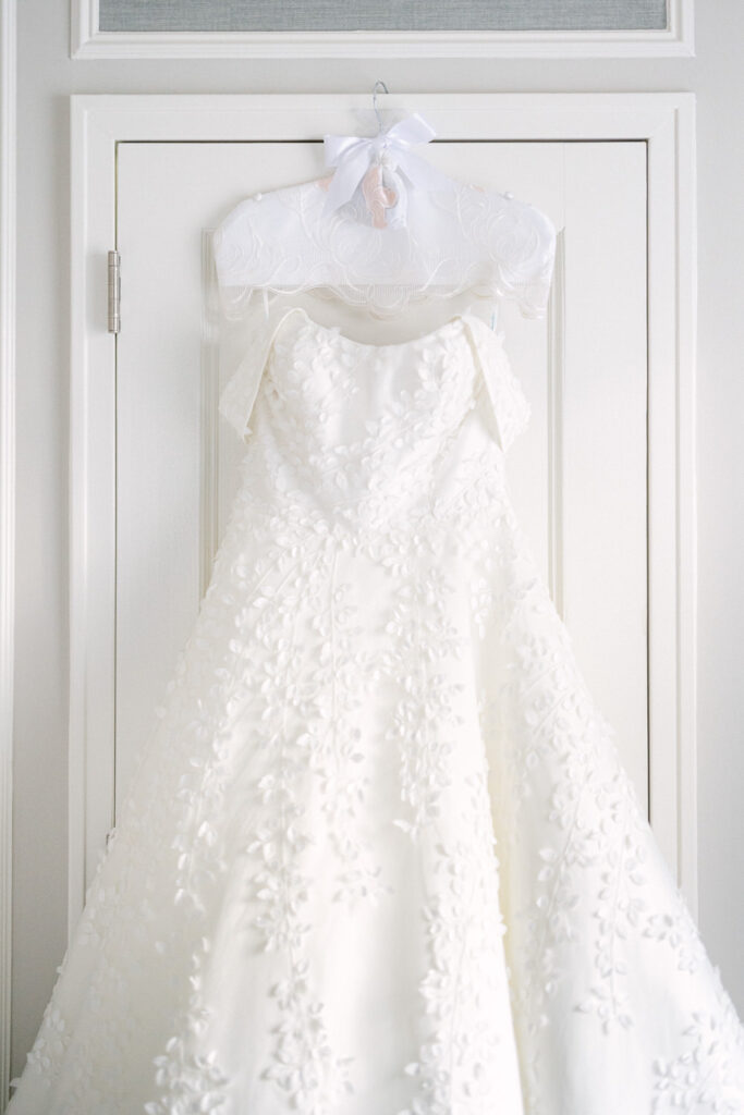 A beautiful white wedding dress with intricate floral embroidery, displayed on a hanger against a light-colored door.