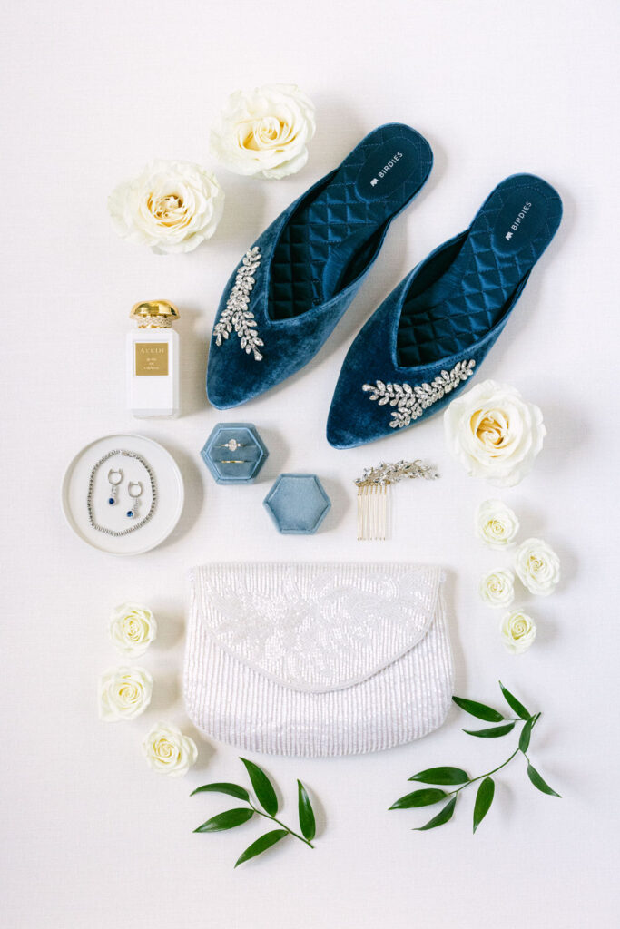 A flat lay arrangement of stylish blue velvet shoes adorned with embellishments, a white clutch bag, delicate floral accents, jewelry pieces, and a perfume bottle, all set on a light textured background.