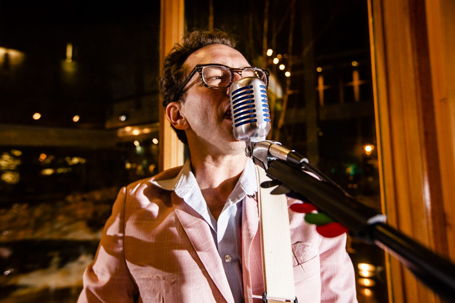 A musician passionately singing into a vintage-style microphone, wearing a pink blazer, with a warm, inviting atmosphere illuminated by soft lights.