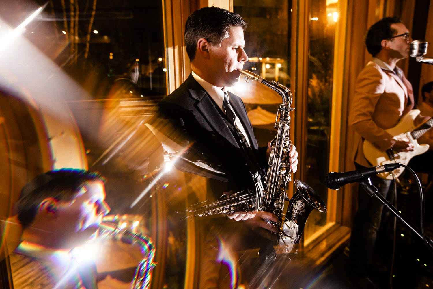 A saxophonist passionately plays on stage, illuminated by soft lights, with another musician performing in the background.