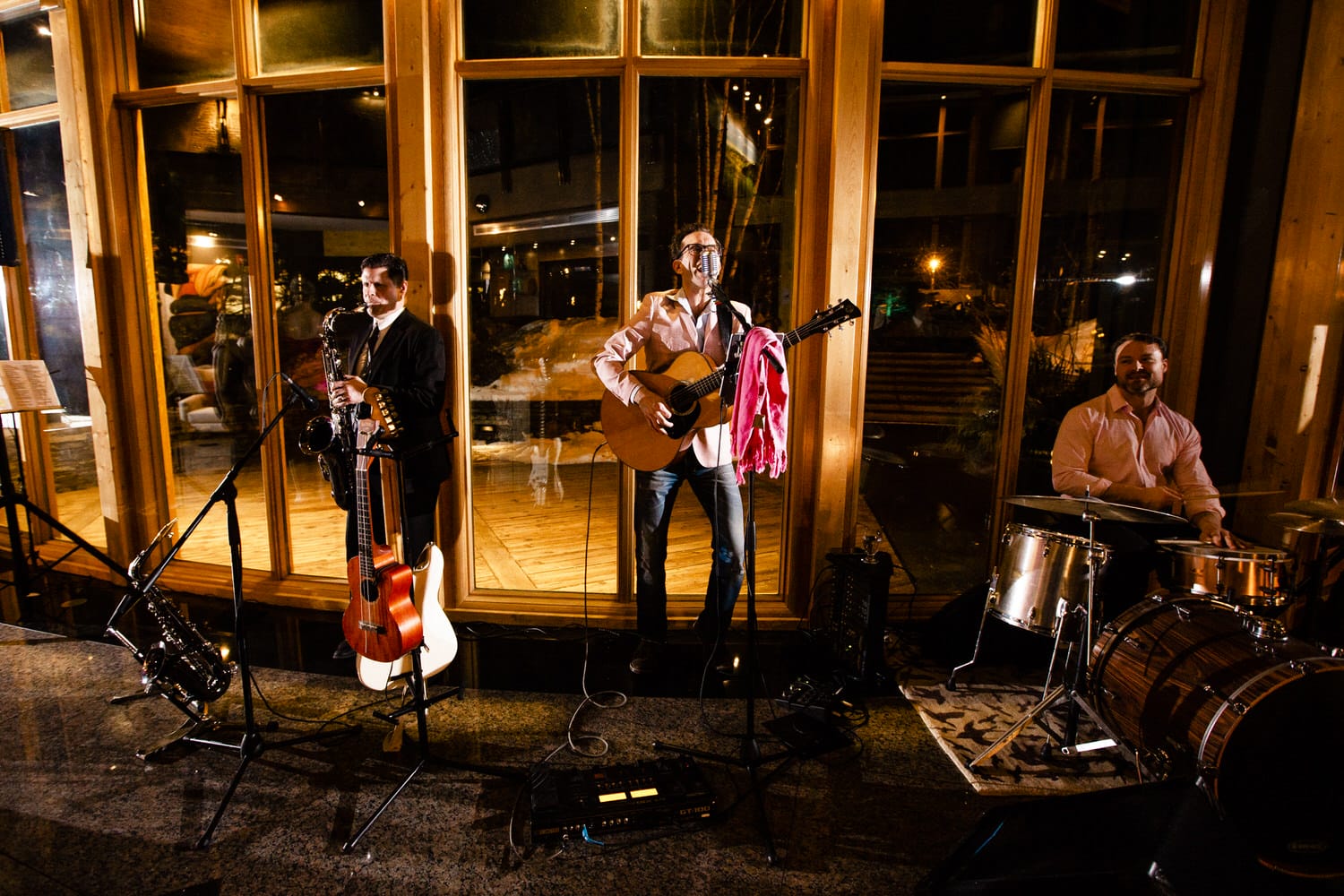 A band performs live with instruments, including a guitar, saxophone, and drums, in a warmly lit space with large windows showing a nighttime view.